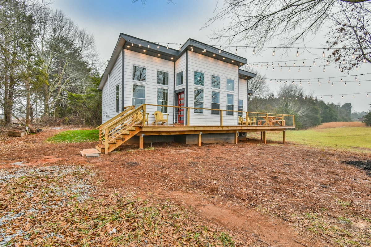 A modern home with large windows is presented, featuring a spacious front deck with wooden railings. String lights are visible, providing ambient lighting. Surrounding the house is a natural area with fallen leaves, adding to the outdoor setting.