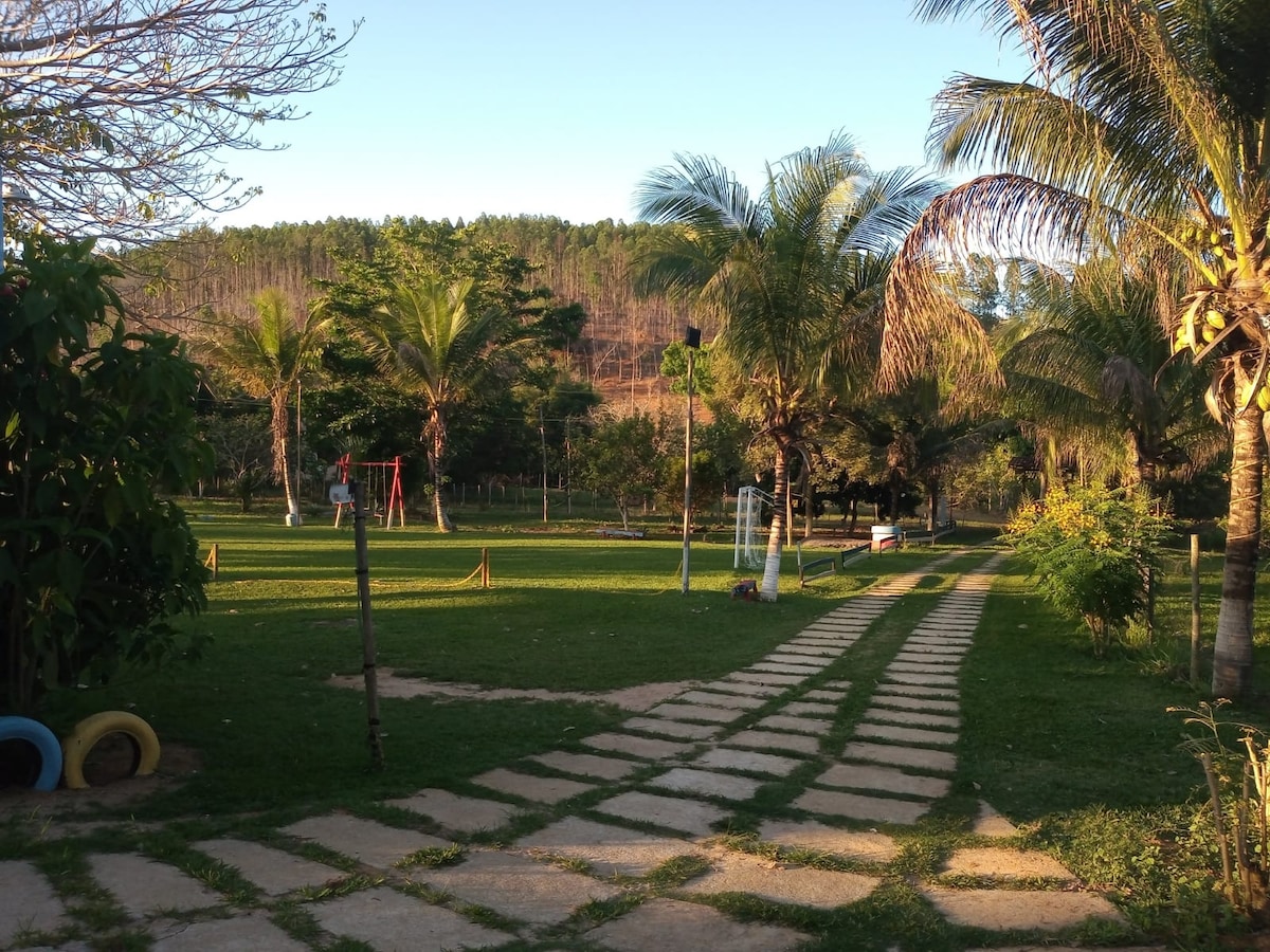 A winding stone pathway leads through a lush green lawn, bordered by palm trees and other foliage. The area features playground equipment visible in the distance, with gentle hills rising in the background under a clear sky.