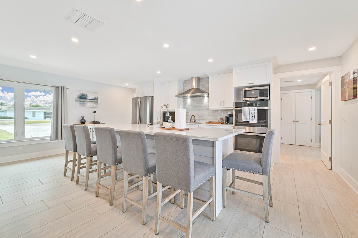 A modern kitchen features a large island with seating for eight, complemented by light gray upholstered chairs. Stainless steel appliances are visible, along with white cabinetry and a range hood. Natural light floods in from large windows, highlighting the open floor plan.