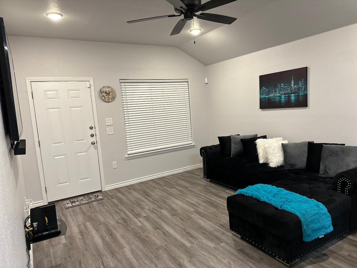 A cozy living area is equipped with a dark sectional sofa adorned with decorative pillows. A vibrant blue throw adds a pop of color. A ceiling fan is present above, and natural light enters through a window with horizontal blinds, while a flat-screen television is mounted on the wall.