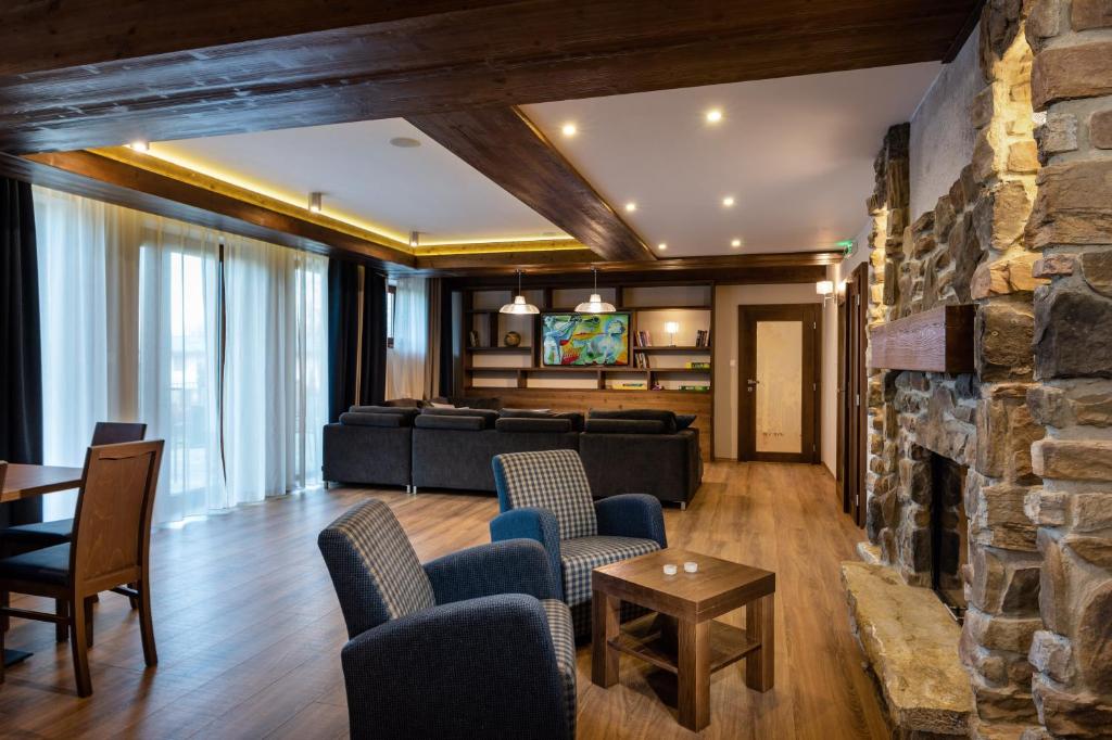 A spacious living area features wooden beams in the ceiling and a stone fireplace. Comfortable seating is arranged around a coffee table, with large windows allowing natural light to fill the space. A dining table sits nearby, complementing the warm wood tones of the room.