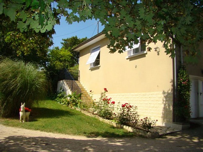 The exterior of a quaint house is viewed, complete with a raised entrance accessed by a few steps. Lush greenery surrounds the property, with a mix of flowers and bushes framing the path, providing a welcoming entryway.