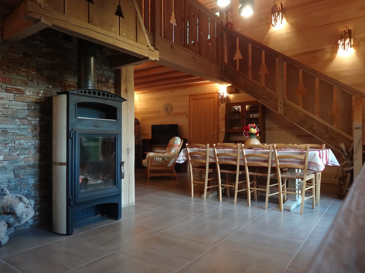 A cozy living space features a stone wood stove and wooden staircase leading to the upper floor. A dining table with several wooden chairs is positioned next to a seating area, with a TV visible in the background. Warm wood accents enhance the cabin's charm.
