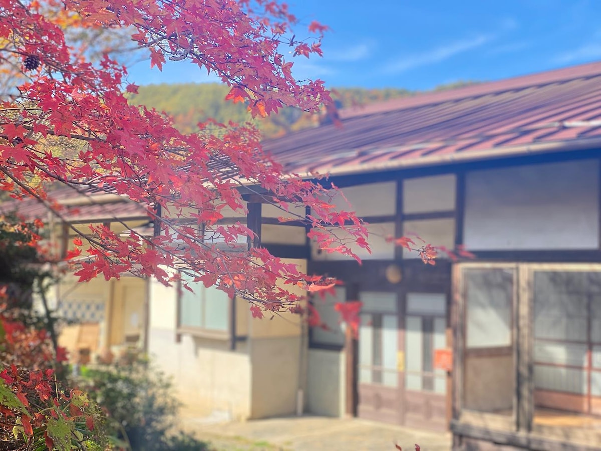 A traditional-style building is framed by vibrant red maple leaves, showcasing a welcoming entrance with sliding doors. The rustic exterior reflects a blend of nature and architecture, with large windows allowing ample natural light to illuminate the space.