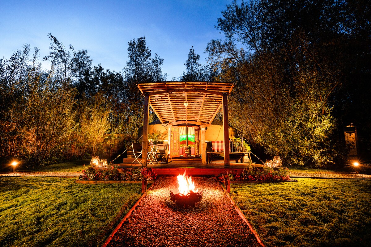 The exterior of Whittlers Yurt is illuminated at dusk, featuring a spacious deck covered by a rustic roof. A fire pit is positioned in the foreground, surrounded by landscaping, while soft lighting enhances the entrance, inviting relaxation in a serene natural setting.