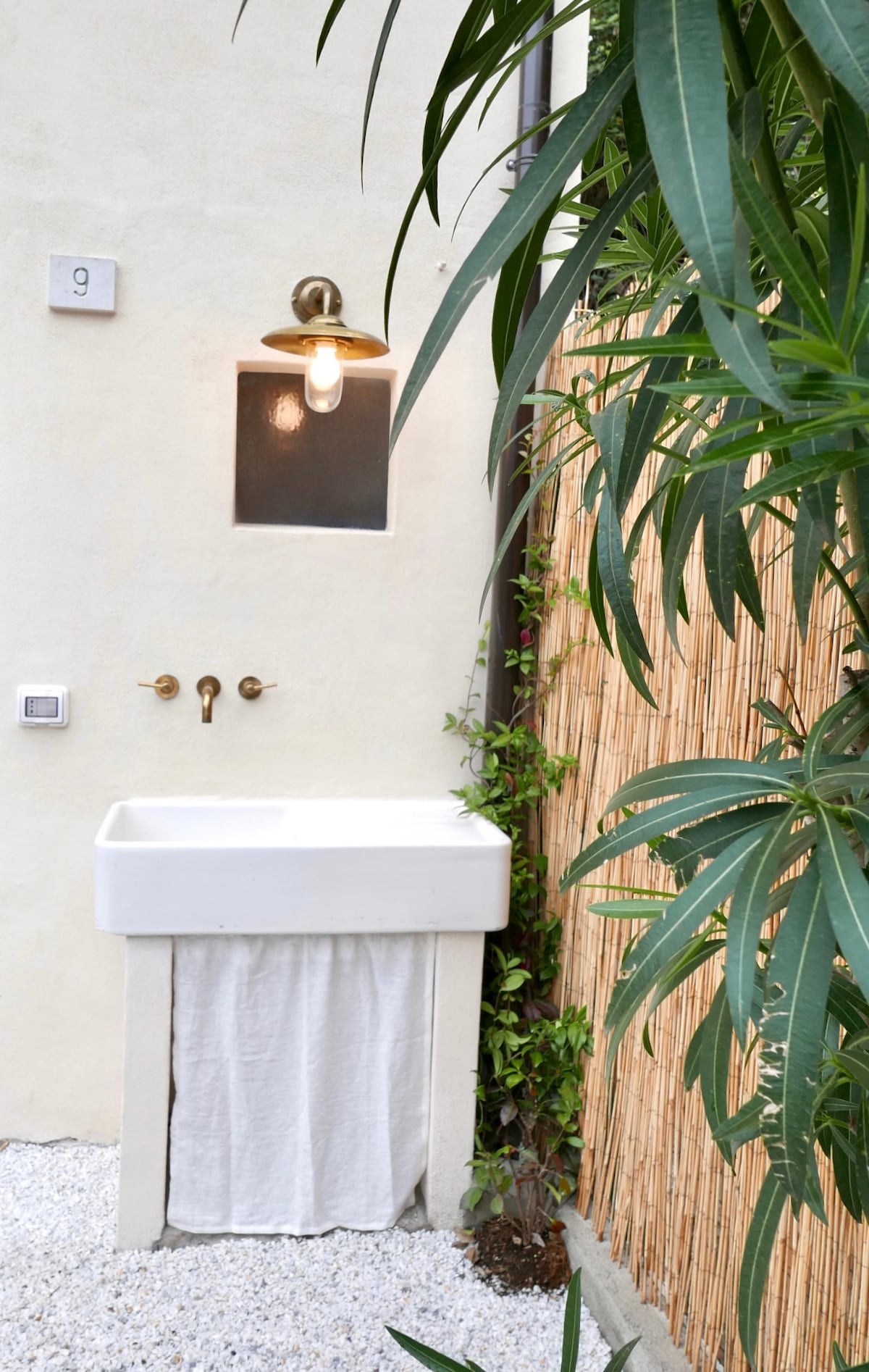 An outdoor garden sink is positioned against a beige wall, featuring brass fixtures and a light fixture above. A soft towel hangs from the front of the sink. Lush green plants surround the area, adding natural elements to the garden space.