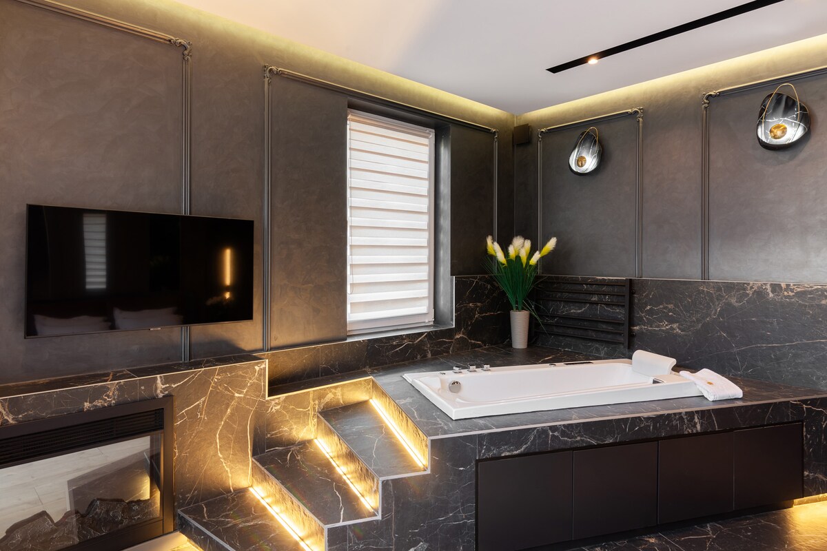 A modern bathroom features a jacuzzi tub surrounded by dark marble walls. Soft lighting highlights the steps leading to the tub, while a television is mounted above a sleek black cabinet. Minimalist decor includes a vase with fresh flowers, adding a touch of color.