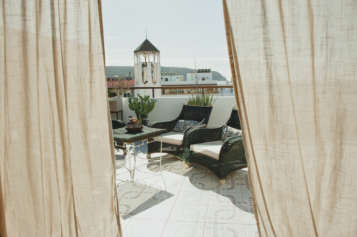 A spacious terrace is visible, framed by flowing curtains. Comfortable seating arrangements with lounge chairs and a table are positioned on a patterned tiled floor. Lush greenery and a gazebo-style structure add charm to the outdoor space, with distant hills visible in the background.