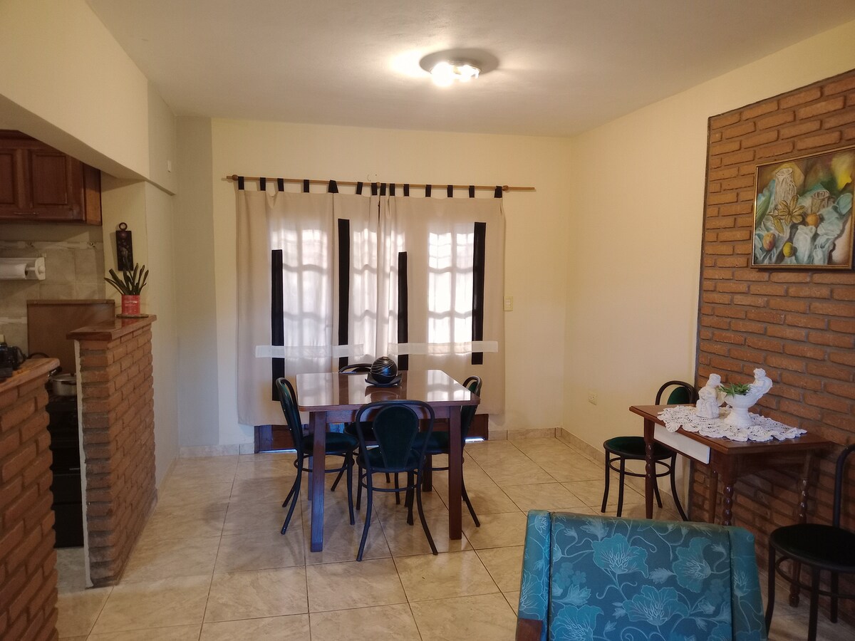 A bright dining area is seen, featuring a wooden table surrounded by four dark green chairs. Natural light filters through sheer curtains over two windows, complemented by a textured brick wall. A cozy seating arrangement is present in the corner.