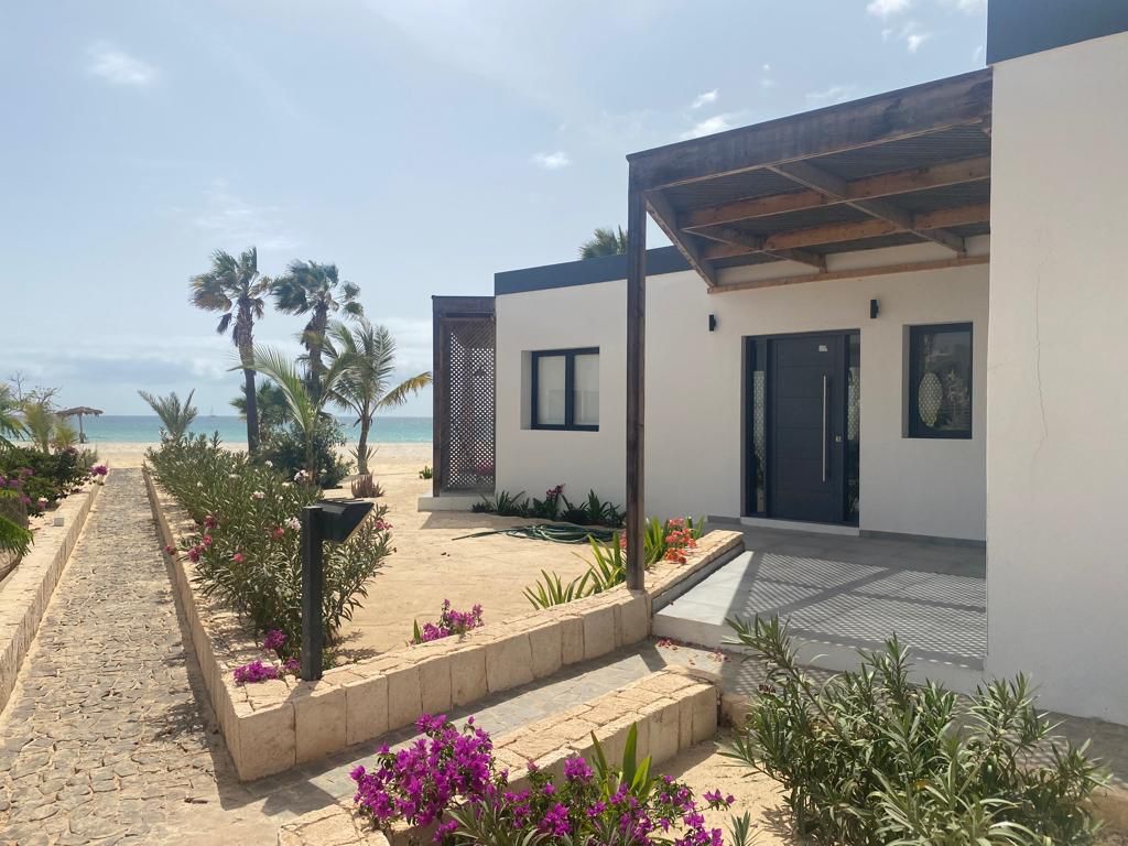 A modern exterior view of the house showcases a welcoming entrance framed by lush greenery and vibrant flowers. The path leads to the door, with a backdrop of swaying palm trees and a visible stretch of beach in the distance.