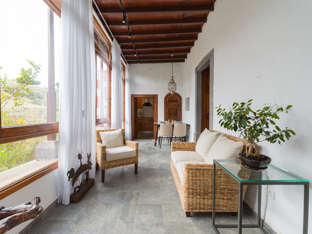 A sunlit seating area features natural wicker furniture, including a sofa and a glass side table. Large windows allow views of the surrounding landscape, while white curtains softly diffuse the light. A dining area is visible in the background with wooden accents.