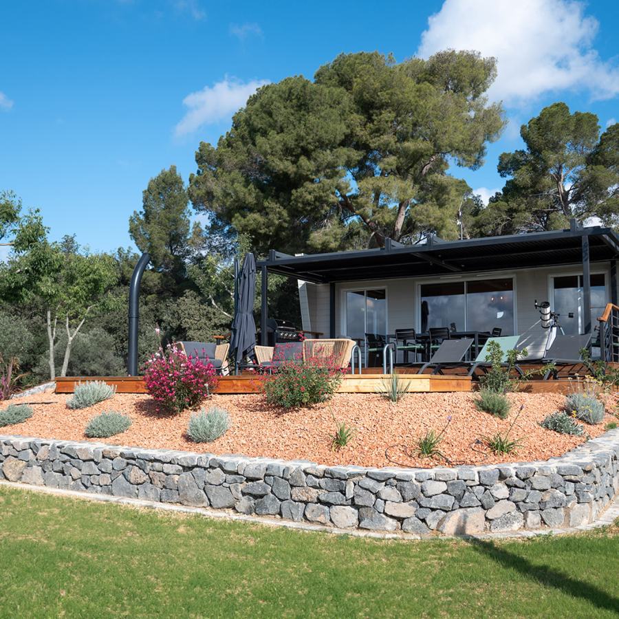 A modern cottage is set against a backdrop of trees, featuring a wooden deck with seating and a dining table. Surrounding gardens are adorned with various plants and shrubs, while the pool area is highlighted by shaded umbrellas. An astronomical telescope is positioned for night sky observation.