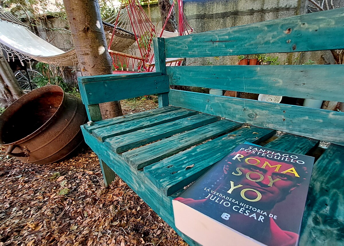A wooden bench in a garden setting is adorned with a large book featuring a colorful cover. Surrounding elements include a hammock in the background and a large, rustic pot nearby. Leaves cover the ground, creating a natural and inviting atmosphere.