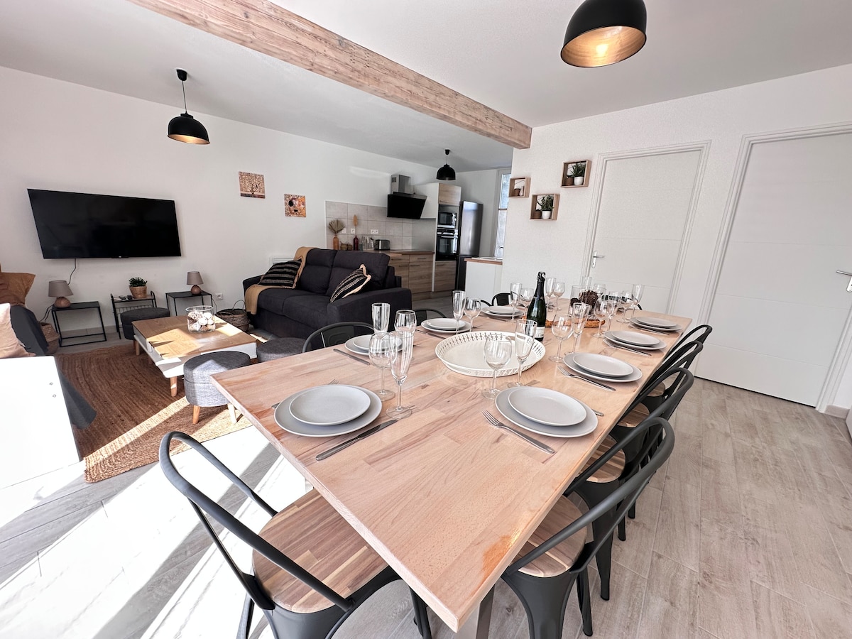 A spacious dining area features a large wooden table set for eight, with plates and glasses arranged neatly. Behind, a well-lit living area includes a comfortable sofa and a flat-screen TV mounted on the wall, creating an inviting space for gatherings.