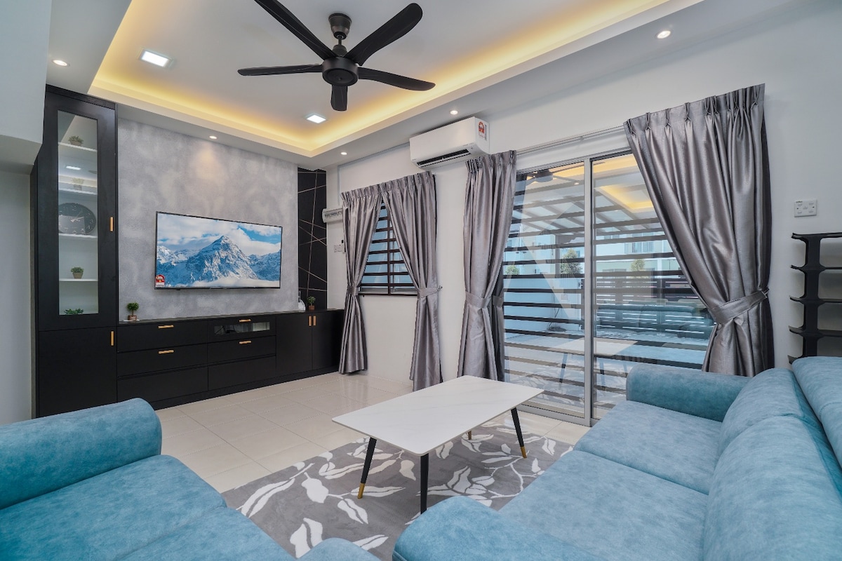 The living room features a comfortable blue sofa set arranged around a low coffee table. A large flat-screen TV is mounted on a feature wall, and light filters through expansive glass doors, complemented by elegant curtains. Soft ceiling lighting enhances the space.