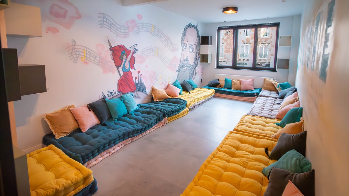 A spacious seating area features large, colorful floor cushions arranged in a U-shape. The wall is decorated with a whimsical mural of musical notes and artistic figures. Natural light enters through large windows, providing a bright atmosphere.