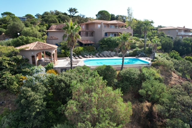 An aerial view captures a spacious property nestled among lush greenery, featuring a shared infinity pool surrounded by sun loungers. A shaded gazebo provides a relaxing spot near the pool, while the home is set against a backdrop of trees and gentle slopes.