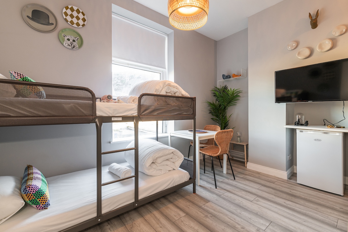 The studio features a bunk bed positioned by a large window, allowing natural light to fill the space. A small dining area is present, complete with a table and two chairs. A wall-mounted TV and a mini-fridge are also visible, providing convenience for guests.