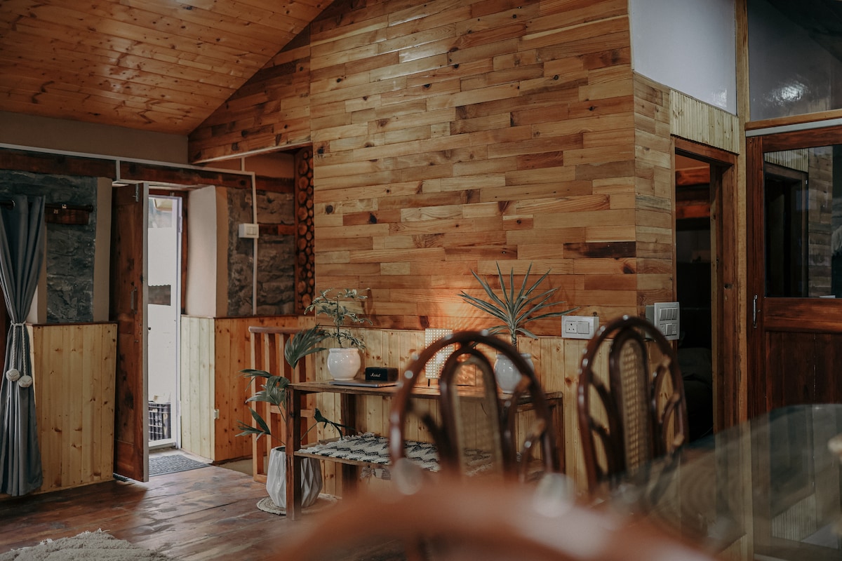 The interior features warm wooden walls and ceilings, contributing to a cozy atmosphere. A dining area with woven chairs is visible, along with a small table. A door in the background leads to additional spaces, while a potted plant adds a touch of greenery.
