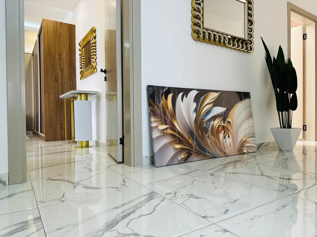A hallway is presented with glossy marble flooring, reflecting light from the surroundings. A large, framed artwork featuring flowing gold and cream designs leans against the wall, while a tall potted plant adds a touch of green. Modern mirrors and wooden accents are visible in the background.