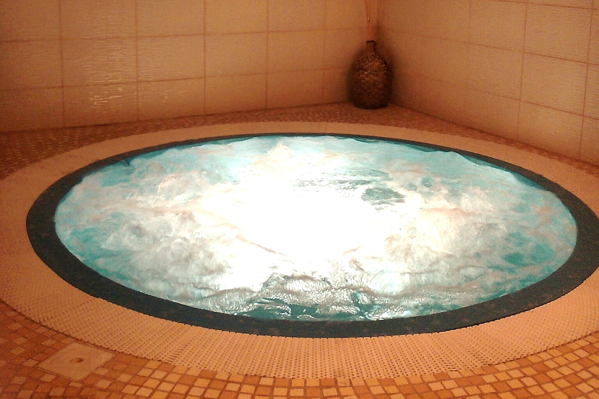 A round spa pool is filled with clear blue water, surrounded by a mosaic-tiled edge. Gentle bubbles can be seen on the water's surface, creating a soothing appearance. A decorative accent piece is placed nearby, contributing to the calming atmosphere.