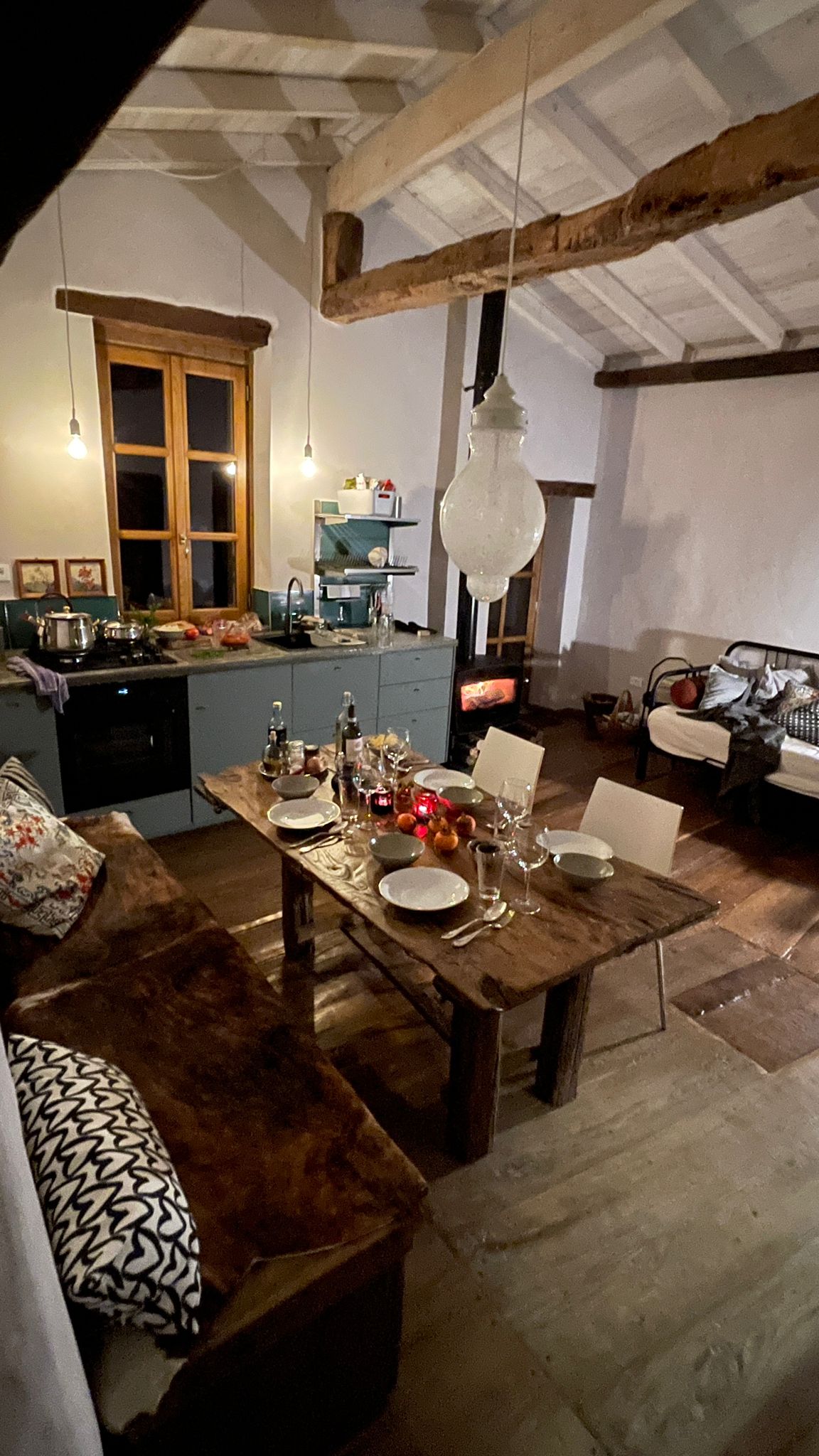 A rustic dining area features a large, weathered wooden table surrounded by white chairs. Warm light from pendant lamps illuminates the space, highlighting the integrated kitchen and cozy seating area with soft furnishings. A wood stove is positioned nearby, adding warmth to the setting.