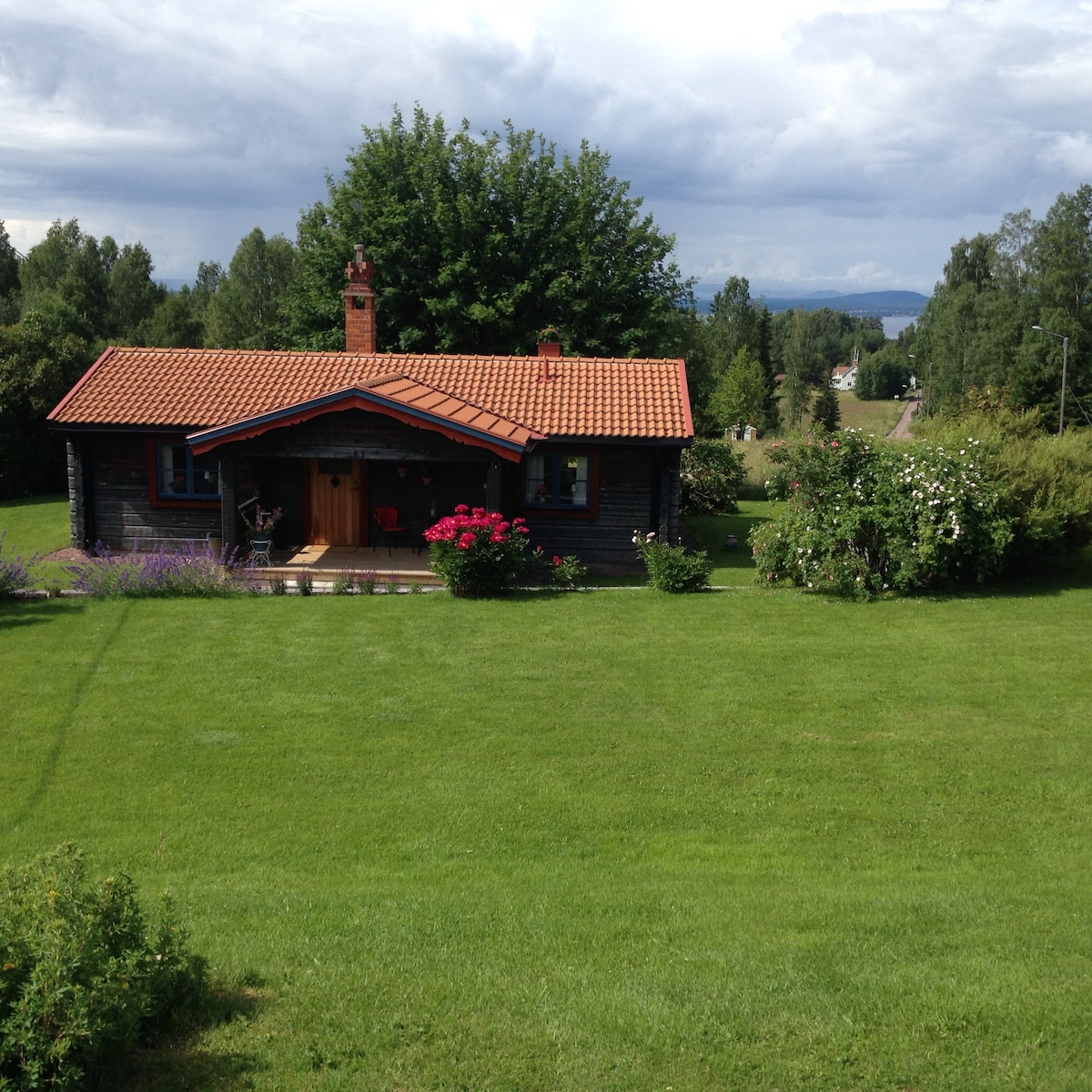 A charming wooden cottage is set on a lush green lawn, surrounded by colorful flower beds. The cozy front porch features a welcoming entryway, while trees and distant hills create a peaceful backdrop under a partly cloudy sky.