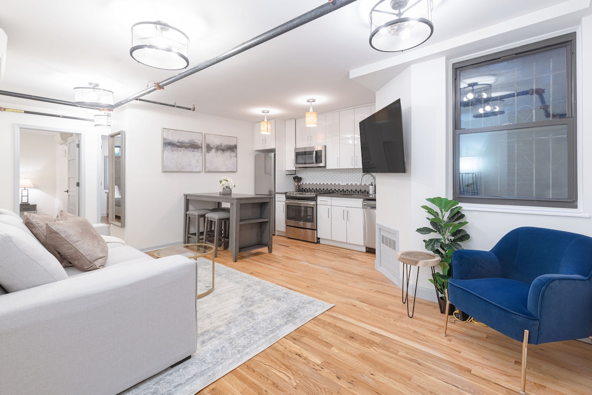 An open living area is presented with a modern kitchen featuring stainless steel appliances and a geometric tile backsplash. A sofa and plush blue chair are positioned on a light wood floor, with a coffee table and decor adding subtle elegance. Natural light enters through large windows.
