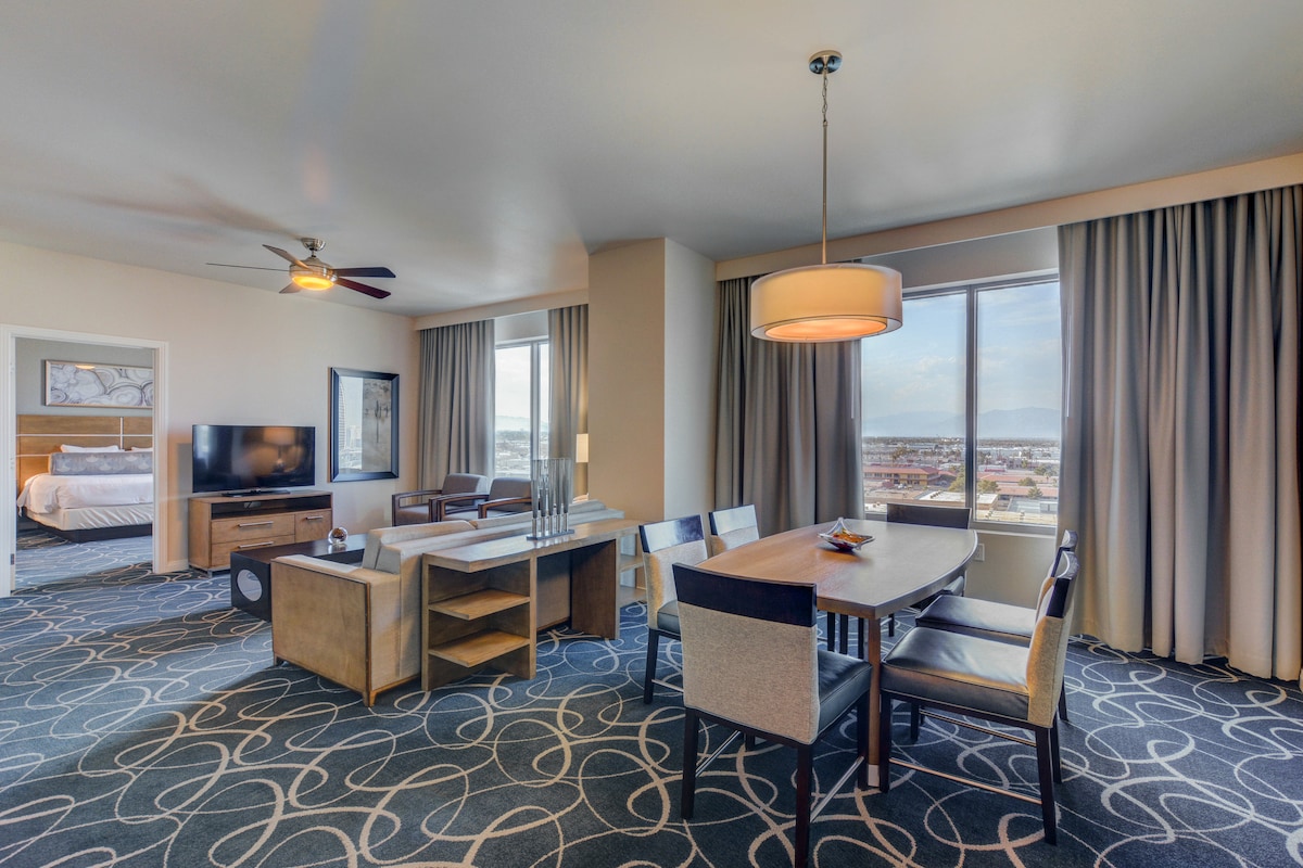 A well-appointed living area features a modern table surrounded by comfortable chairs. A flat-screen TV is mounted on the wall, with a cozy seating arrangement nearby. Natural light fills the space through large windows, complemented by neutral-colored curtains and stylish carpeting.