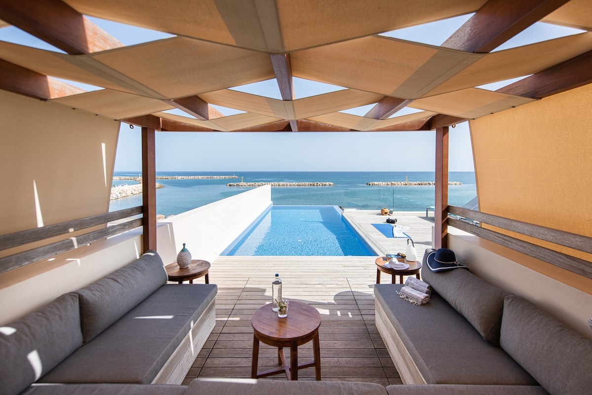 A shaded lounge area is situated on a wooden terrace overlooking the infinity pool and the sea. Comfortable gray sofas are arranged around a small wooden table, showcasing a bottle of water and towels. The stunning views of the water and distant coastline are framed by the pergola.