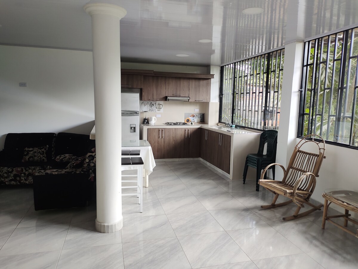 An open kitchen and living area features sleek wooden cabinetry and modern appliances. Natural light is allowed in through large windows. A cozy seating arrangement is highlighted by a patterned sofa and two rustic chairs, complemented by polished tile flooring.