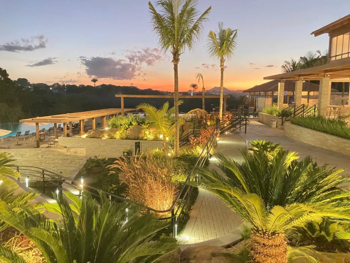 Lush palm trees and vibrant greenery frame a winding pathway leading to a tranquil resort, illuminated by subtle lighting. The warm hues of sunset cast a gentle glow over the landscape, highlighting the outdoor areas and giving a sense of serenity.