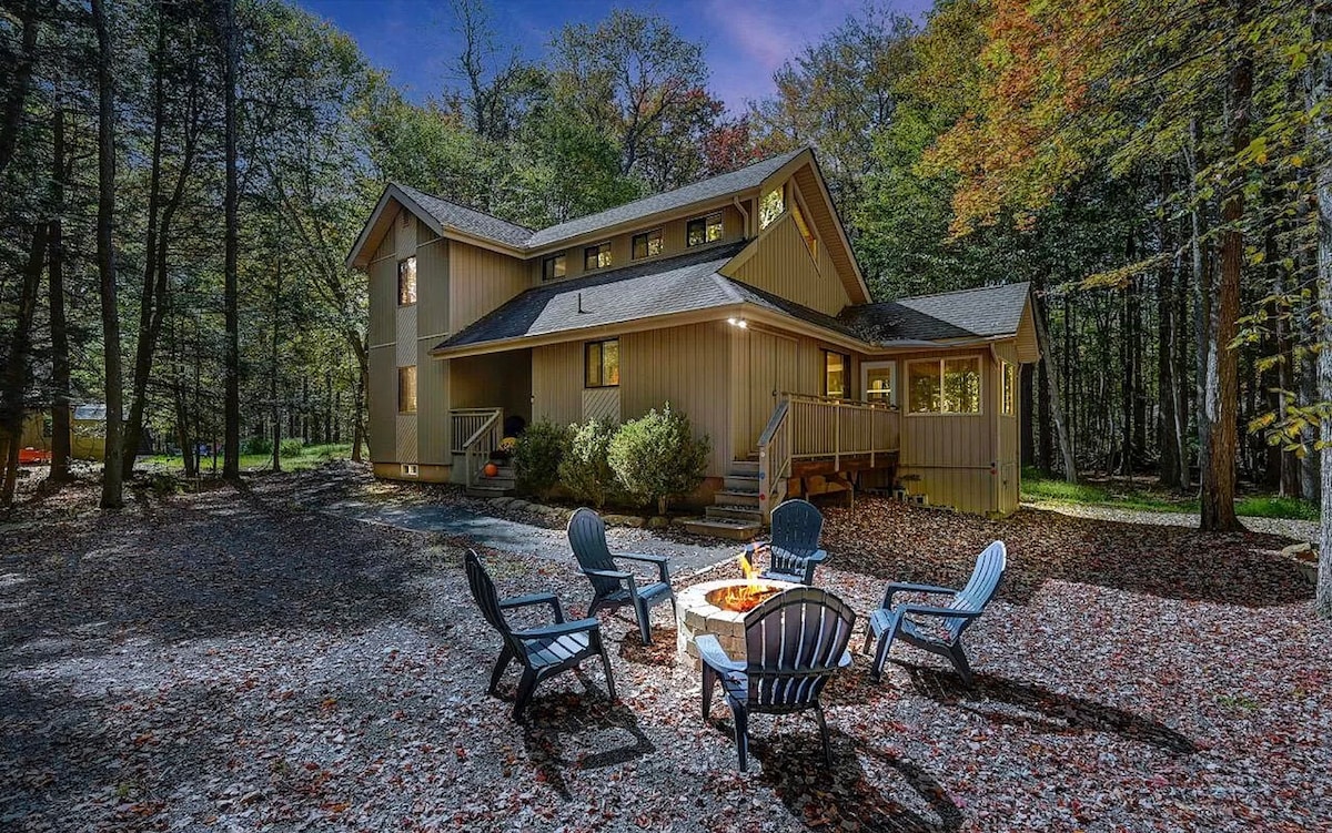 The exterior of a two-story cabin is surrounded by trees, showcasing a wraparound porch and cozy firepit area with adirondack chairs arranged around it. A tranquil atmosphere is created by the soft glow of lights illuminating the home as twilight sets in.