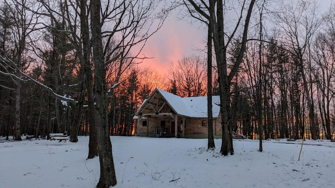 A log cabin chalet is surrounded by snow-covered ground, with tall trees framing the view. The sunset illuminates the sky in shades of orange and pink, creating a serene backdrop for the cozy structure, which showcases a sloped roof and rustic charm.