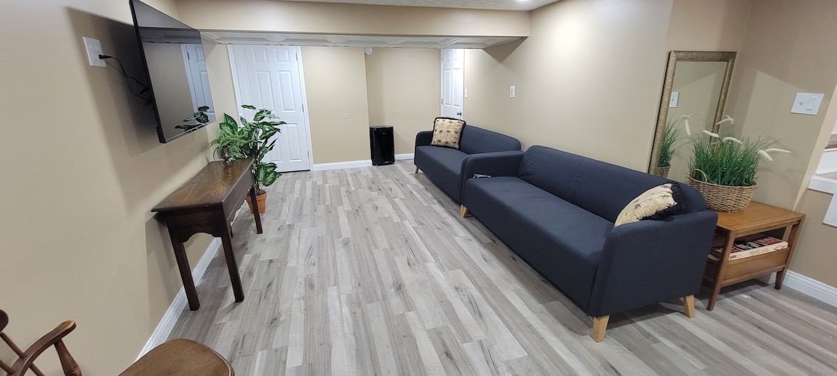 The inviting living area features two modern sofas positioned for comfort. A wall-mounted television casts a focal point, while potted plants add a touch of greenery. The light-colored walls and planked flooring contribute to a spacious, serene environment.