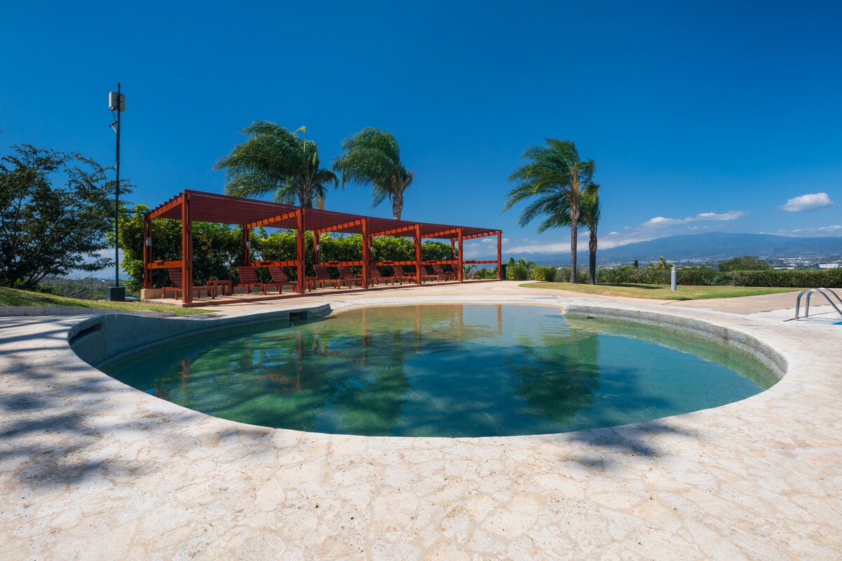 An inviting pool area features a circular pool set in natural stone, surrounded by sun loungers under shaded pergolas. Lush greenery and tall palm trees enhance the tranquil atmosphere, with clear blue skies in the background. The space is designed for relaxation and enjoyment.