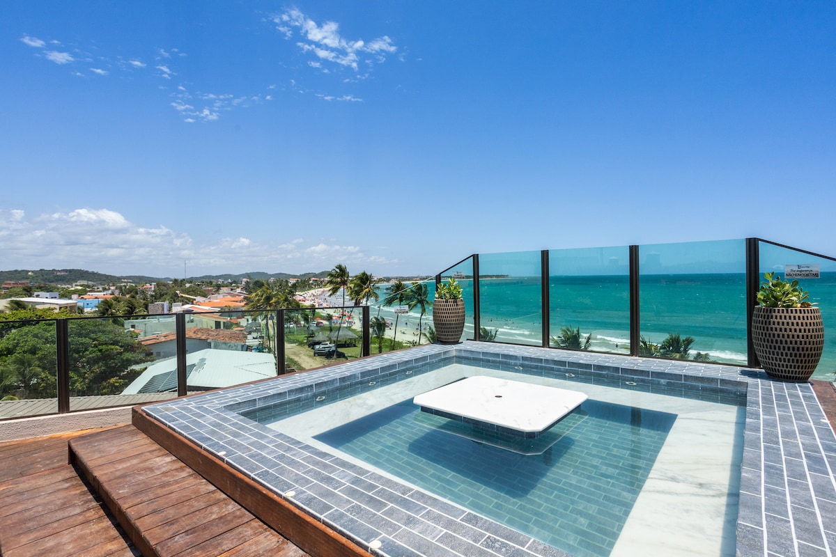 A rooftop jacuzzi is surrounded by glass railings, offering an unobstructed view of the ocean and coastline. The serene blue water contrasts with the natural wood decking below. Potted plants add a touch of greenery, enhancing the inviting atmosphere of this outdoor space.
