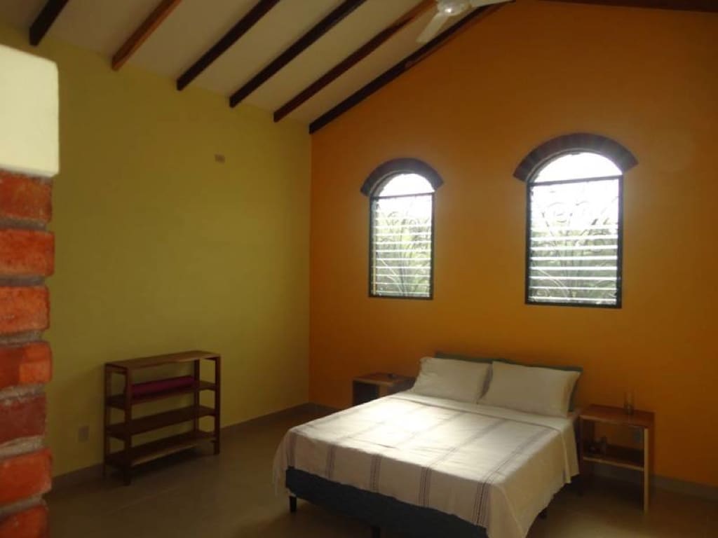 A bright master bedroom features a spacious bed with a simple white blanket. Large arched windows provide natural light, complemented by the warm yellow walls. A wooden shelf is positioned against one wall, with nightstands on either side of the bed.