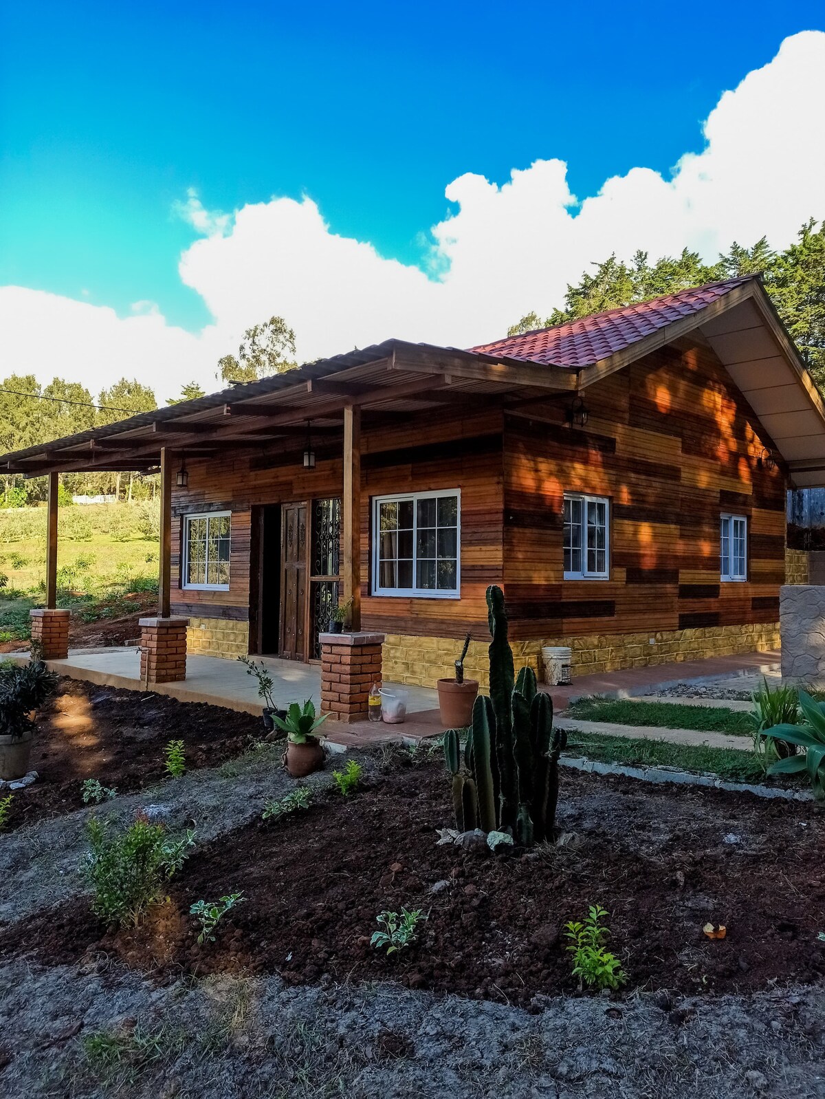 A charming wooden house is surrounded by landscaped gardens featuring various plants, including cacti. The façade is enhanced by stone accents and multiple windows that invite natural light, while a welcoming porch extends along the front, offering a view of the serene landscape.