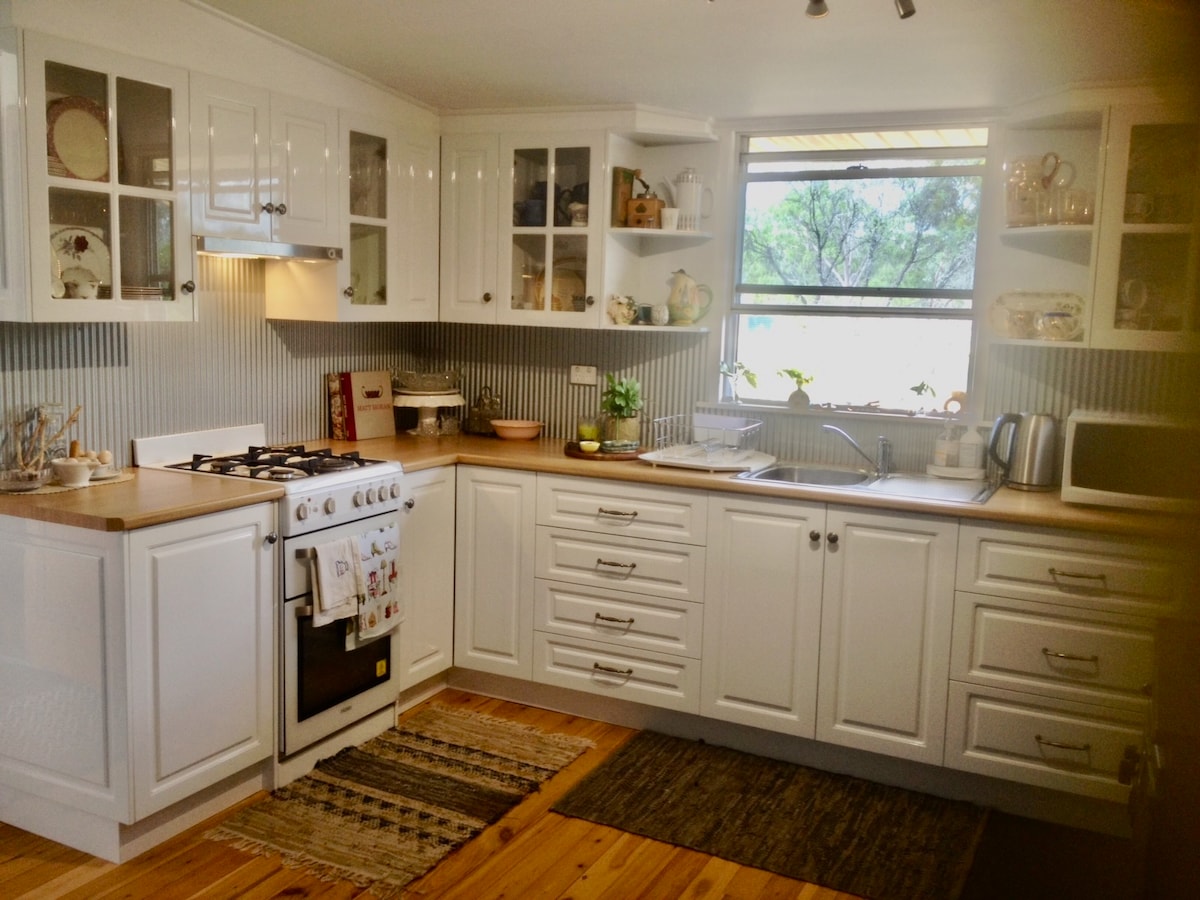 The kitchen features white cabinetry with glass-fronted shelves displaying dishware. A gas stove and a microwave are included among modern appliances, complemented by wooden countertops. Natural light filters through a window, and a woven rug adds warmth to the wooden floor.