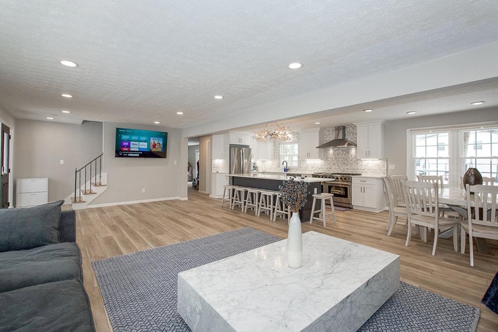 The open living area features a spacious layout with a large marble coffee table at the center. A comfortable seating arrangement is positioned near a wall-mounted television. The modern kitchen, equipped with a chandelier and island, is visible in the background, enhancing the communal atmosphere.