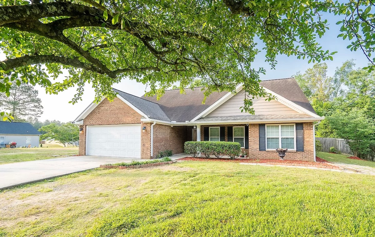 The home features a brick exterior with a welcoming front porch. Fresh green grass surrounds the property, complemented by neatly trimmed bushes. A driveway leads to a garage, and a sizable tree provides shade to the entrance.