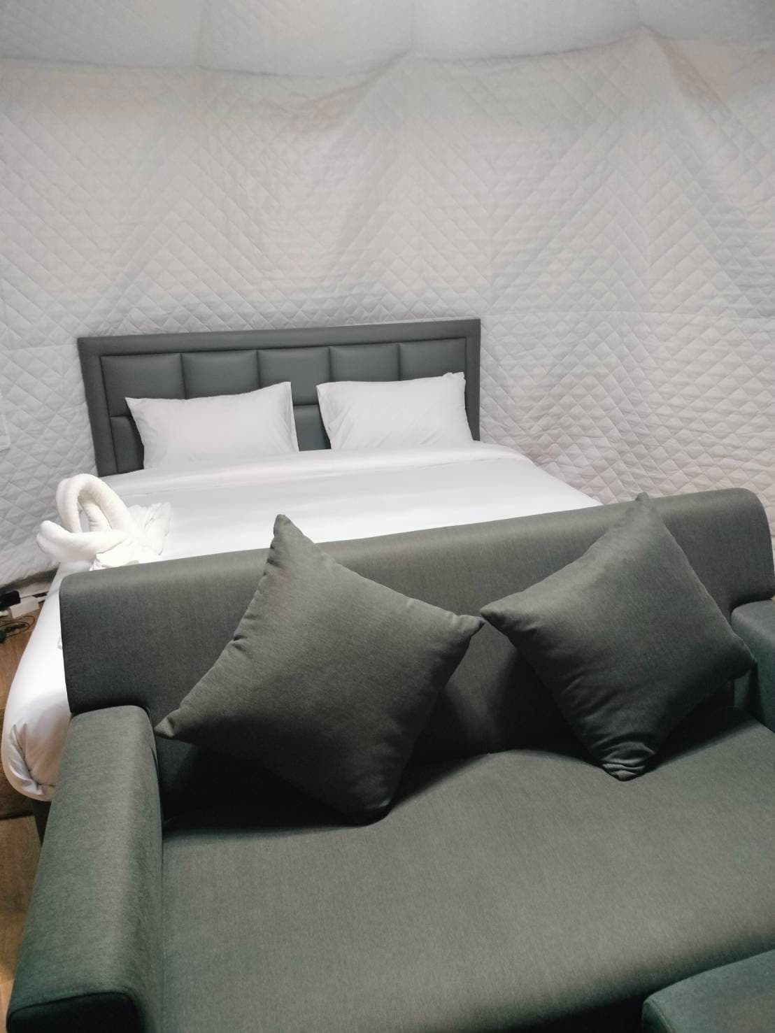 A comfortable sleeping area features a spacious bed with white linens and several pillows. A gray sofa with two accent pillows is positioned near the foot of the bed, surrounded by textured walls draped in white fabric.