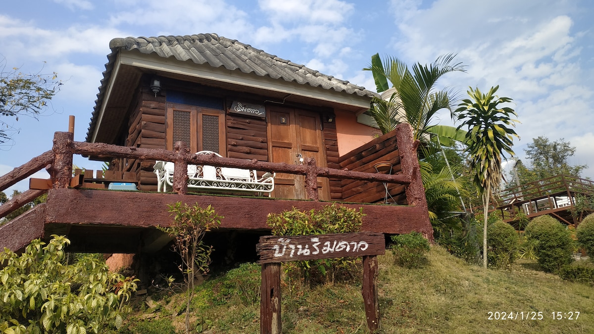A wooden cabin is elevated on a grassy hill, featuring a welcoming front porch with white rocking chairs. Lush tropical plants frame the cabin, and a sign with the property name is prominently displayed in the foreground. The sky is partly cloudy, creating a serene atmosphere.