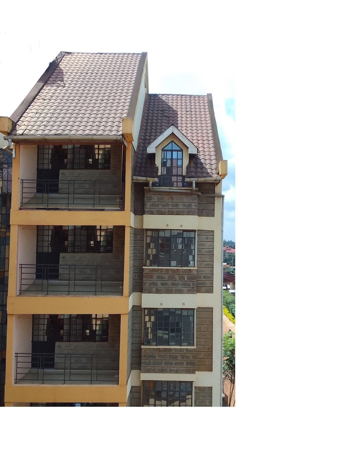 The exterior of a multi-story residential building is displayed, featuring a combination of stone and plaster walls. Symmetrical balconies are present on each level, with black railings providing a modern touch. The roof has a distinctive design, showcasing angled shapes and shades.