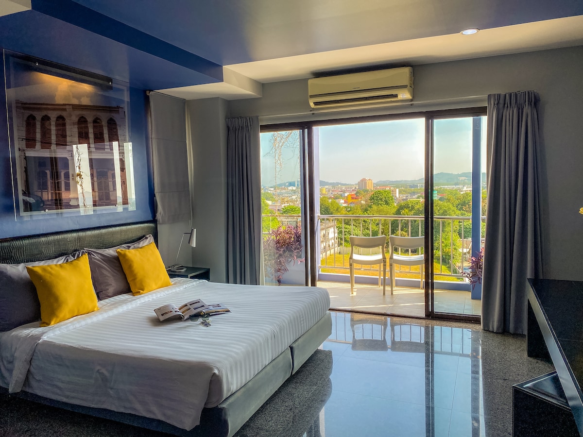 A modern room features a spacious bed with crisp white linens and decorative yellow pillows. A balcony with two chairs offers a view of the surroundings. Large windows allow natural light to fill the space, while air conditioning and a desk add to the comfort.