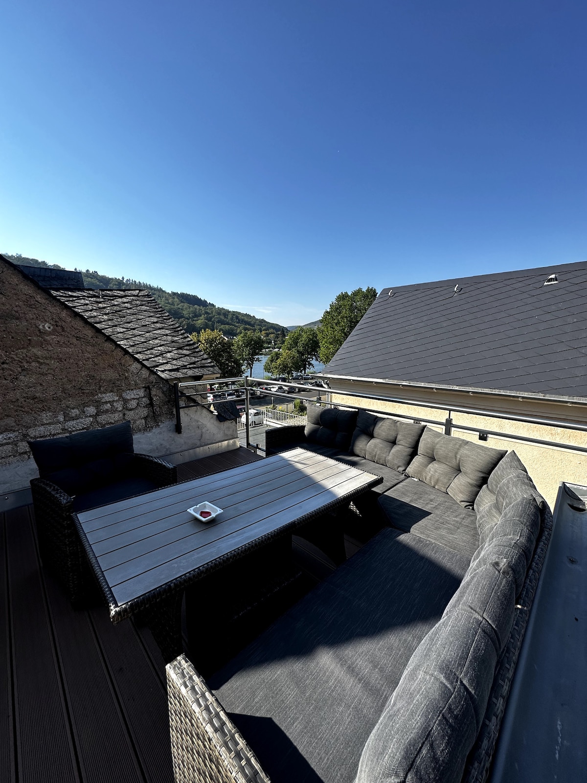 A spacious rooftop terrace is presented, featuring comfortable seating arranged around a table. Views of the surrounding landscape, including hills and the Mosel River, can be observed in the background, with clear blue skies overhead.
