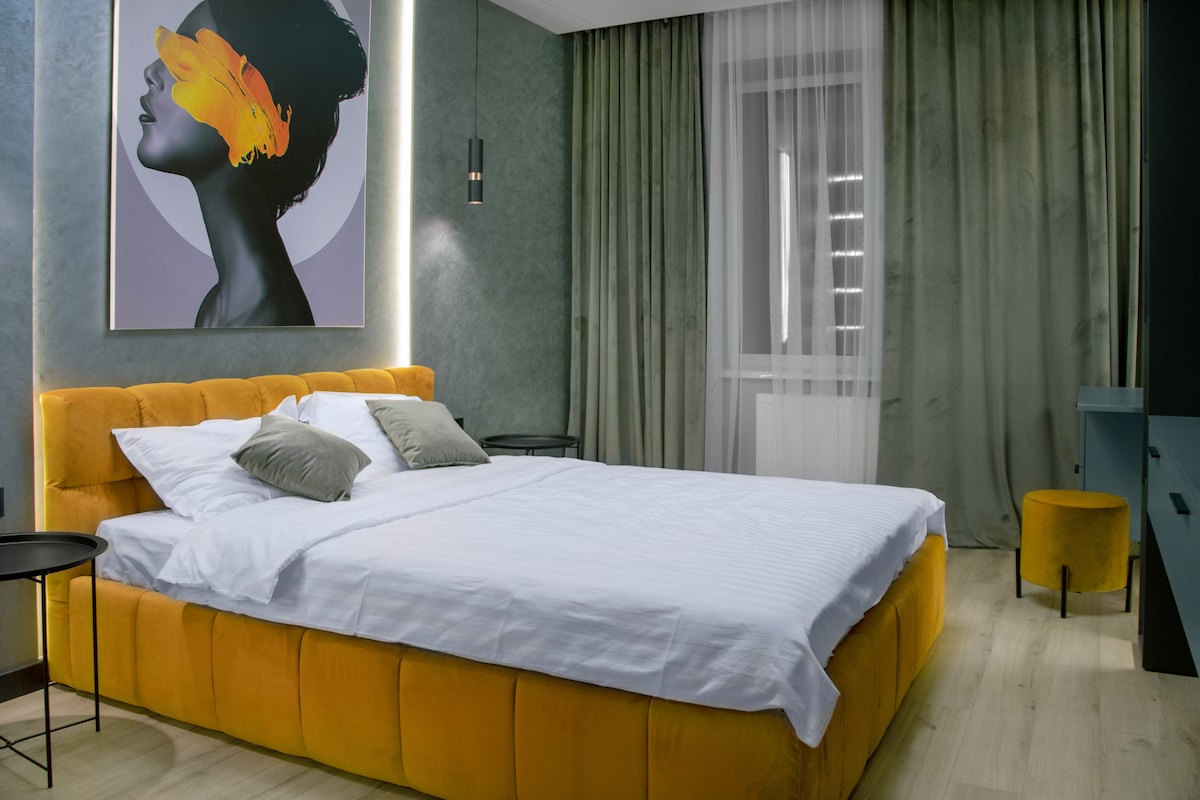A spacious bedroom features a prominent yellow bed with white bedding and decorative pillows. Soft green walls complement the room, while a piece of wall art adds a touch of elegance. Natural light streams in through sheer curtains, enhancing the serene ambiance.