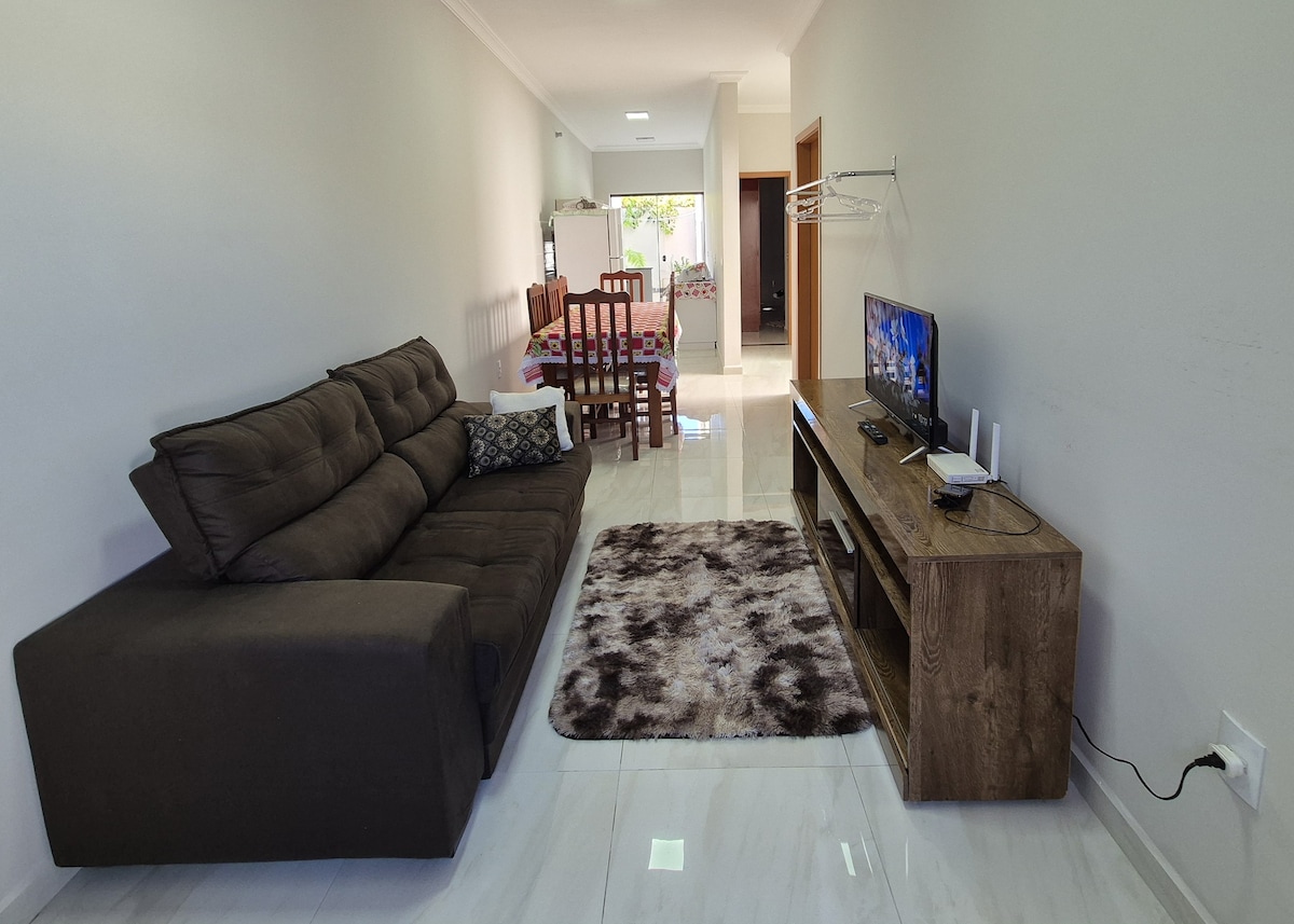 A spacious living area is furnished with a dark sectional sofa and a wooden TV stand. A large rug lies underfoot, and a television is mounted on the stand. The room is well-lit by natural light coming from the adjacent dining area.