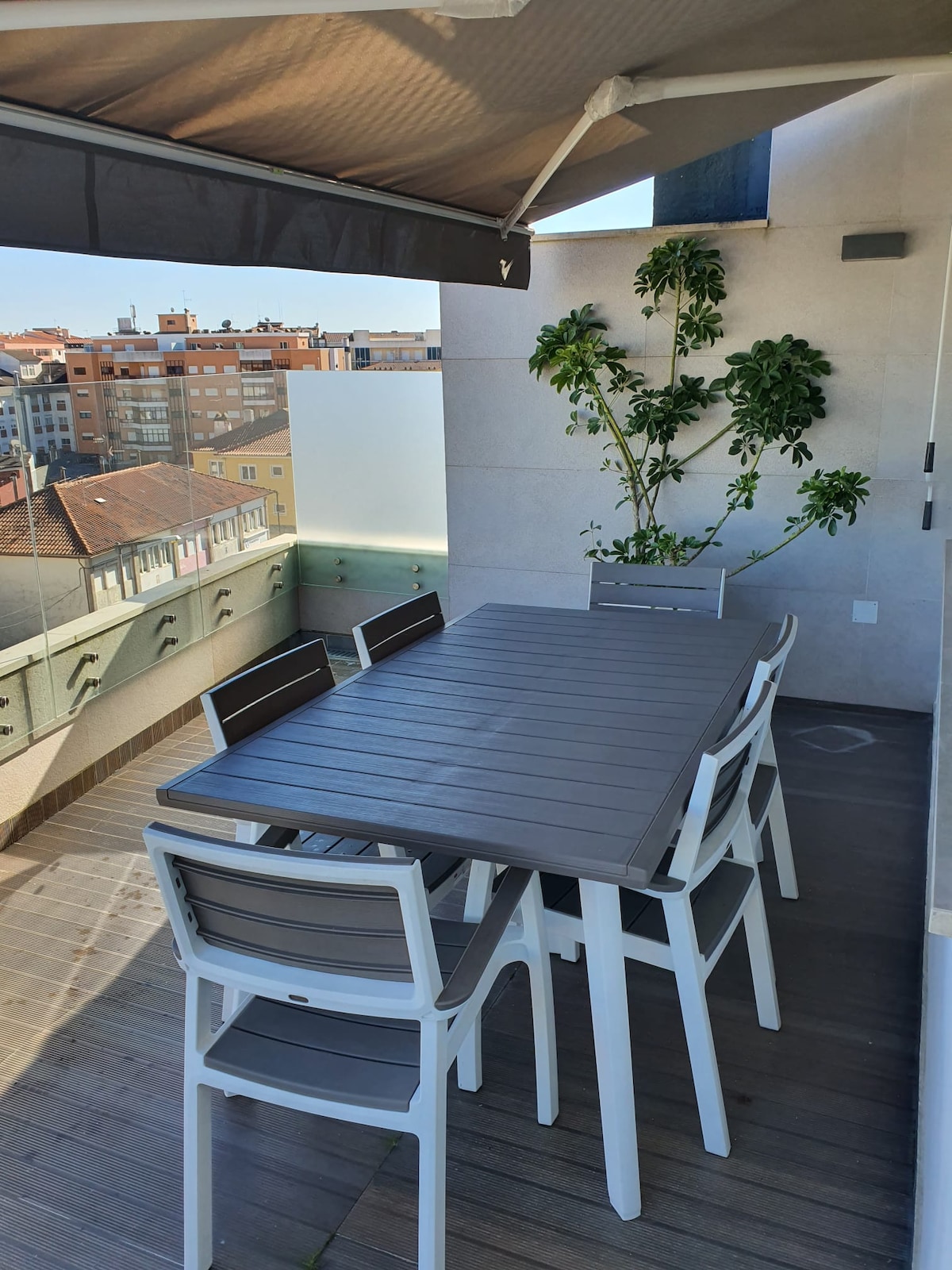 A spacious outdoor balcony features a modern dining table surrounded by six chairs. A small green plant adds a touch of nature. The urban skyline is visible in the background, showcasing a mix of rooftops and buildings under clear skies.